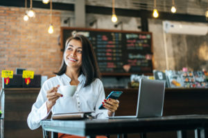 Woman Sitting Happily Working With A Smartphone In A Coffee Shop - Pontual Contadores & Associados